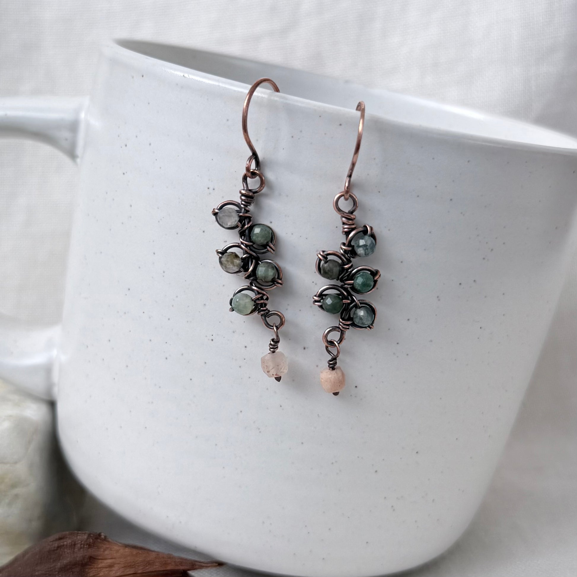 A pair of handmade copper dangle earrings with green gemstone clusters and small peach beads, hanging of a white speckled ceramic mug.