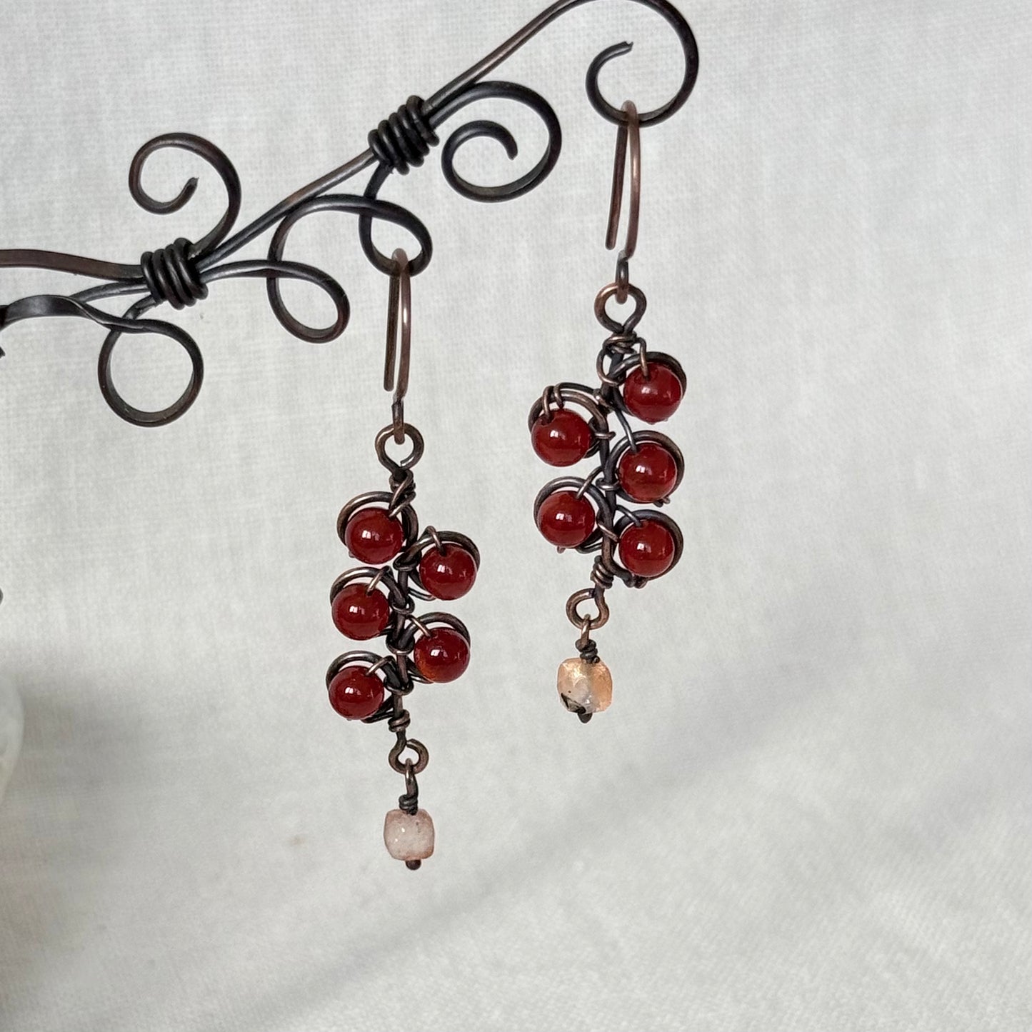 A pair of handmade dangle earrings with copper wire wrapping, featuring clusters of red beads and a small peach stone at the bottom, displayed on a black metal stand against a white fabric background.