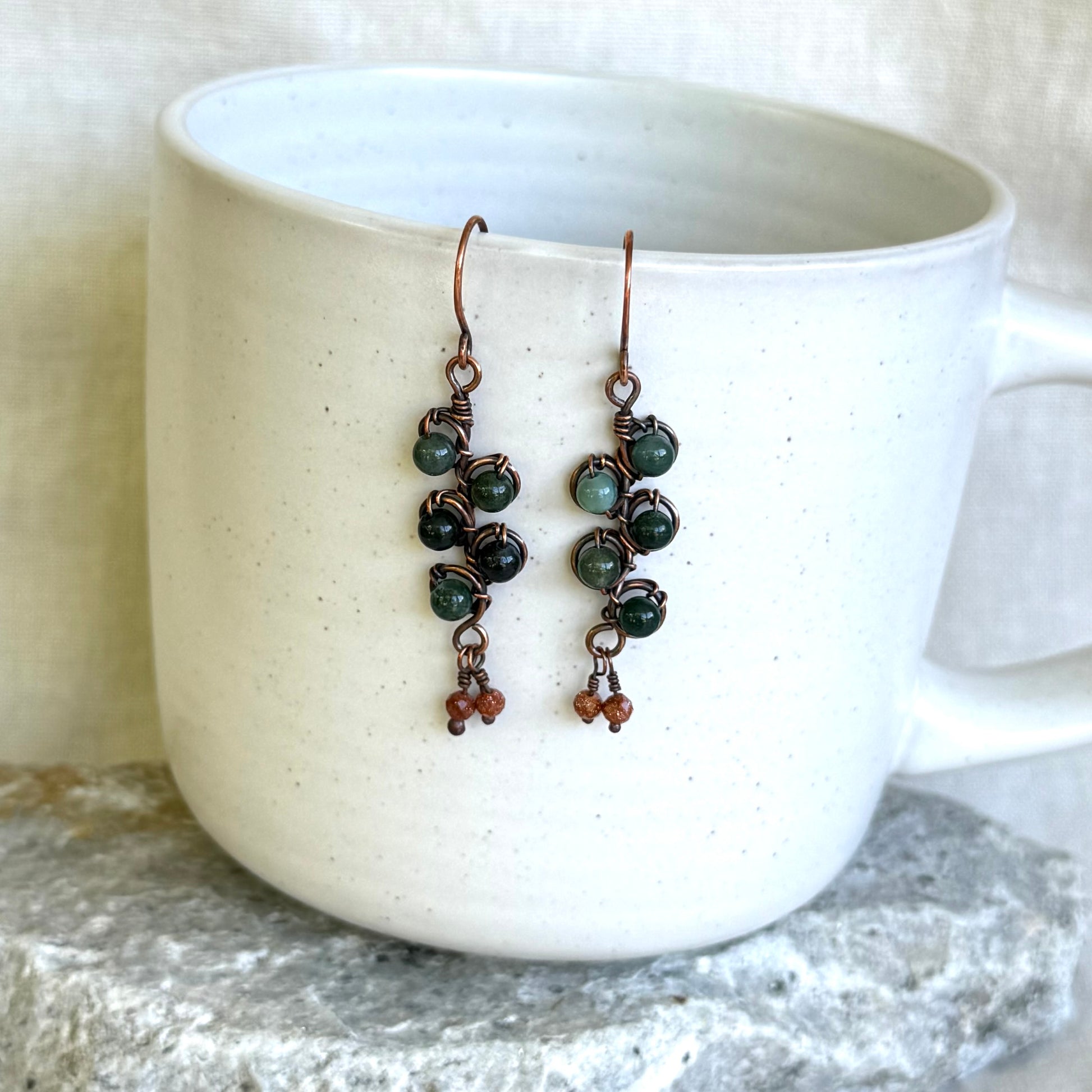 Pair of green beaded copper earrings hanging on a white mug.