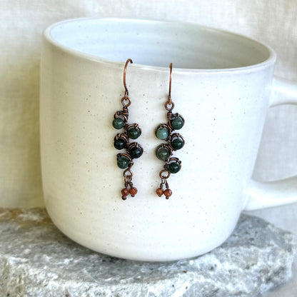 Pair of green beaded copper earrings hanging on a white mug.