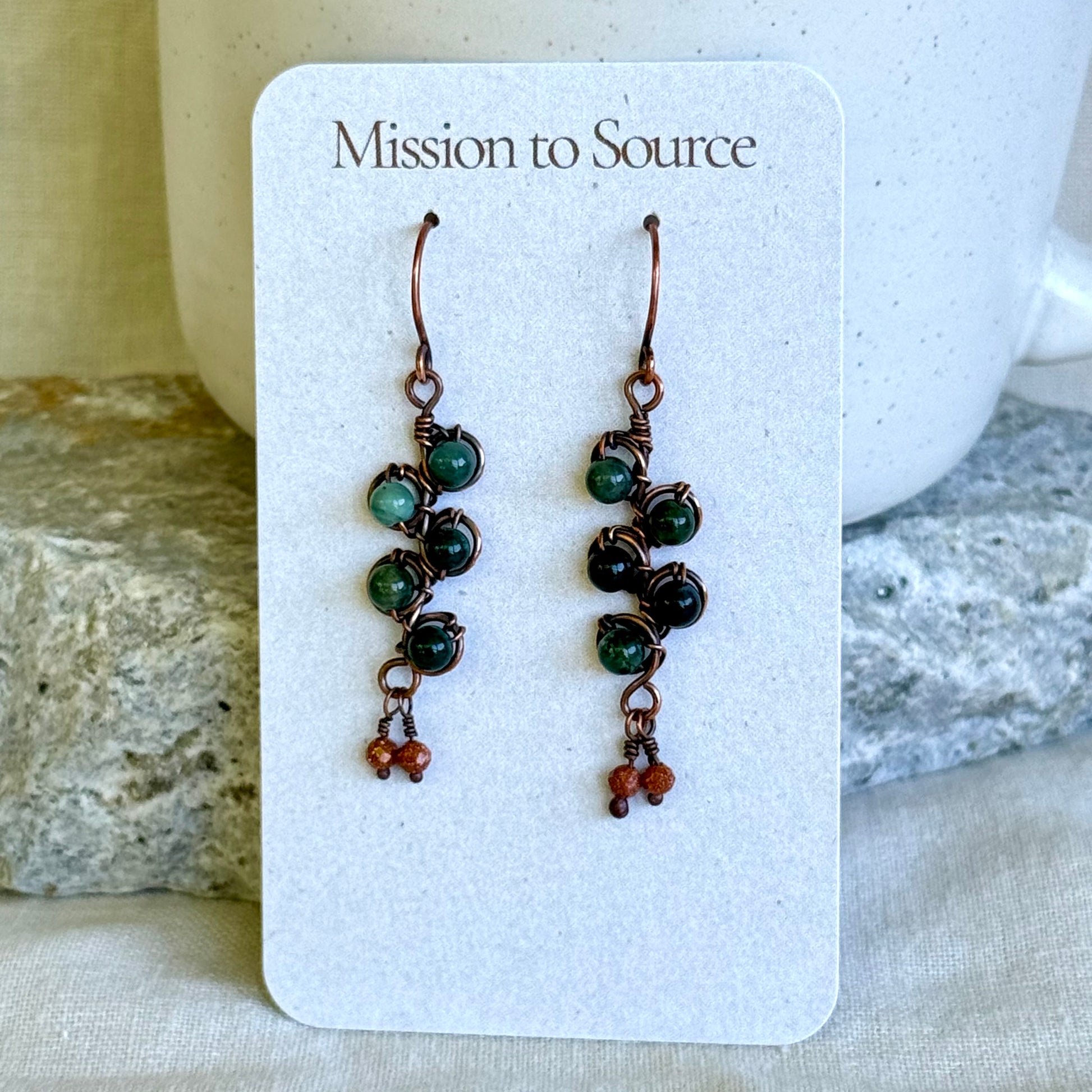 Pair of beaded earrings on a 'Mission to Source' card with a stone and fabric background.