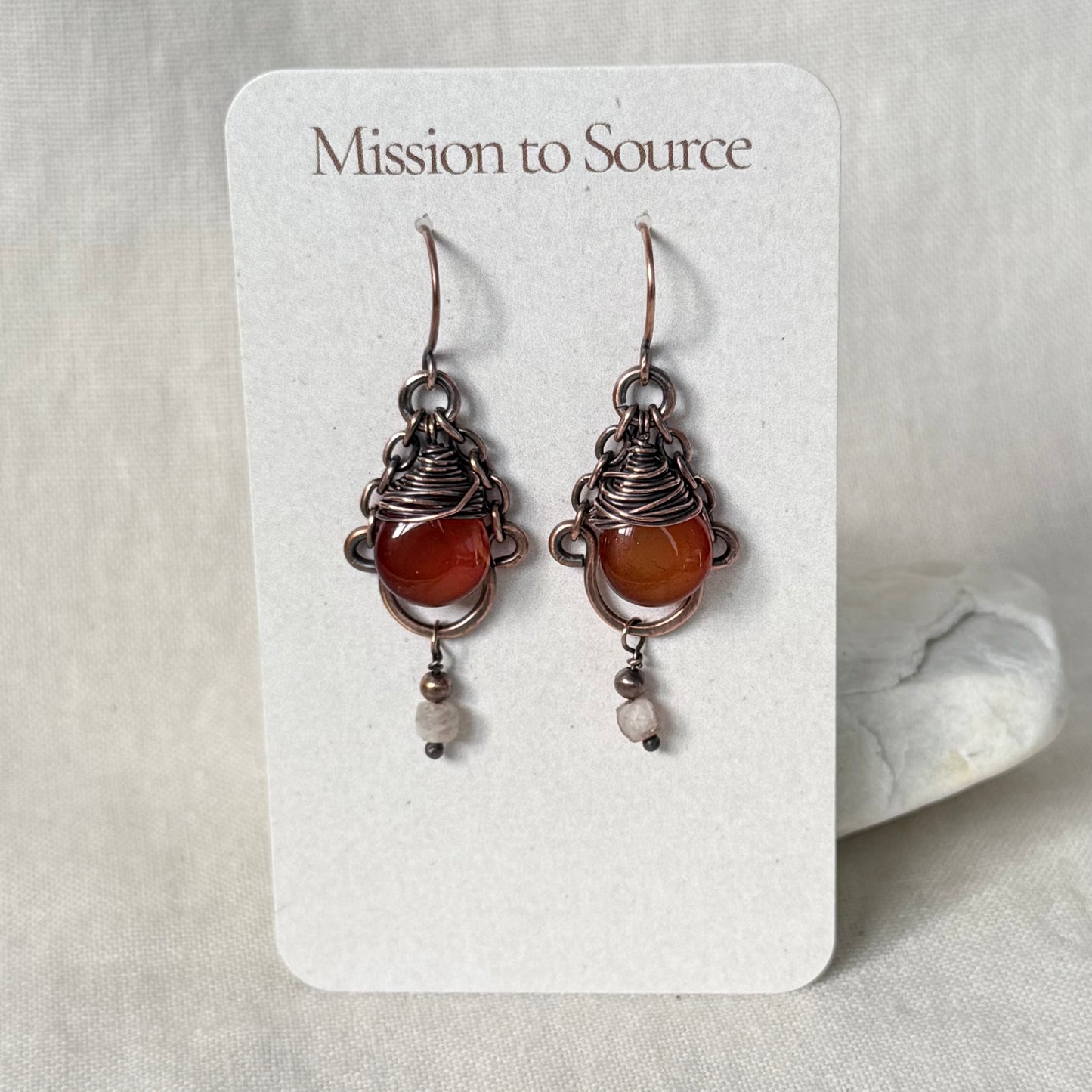 A pair of handmade wire-wrapped earrings with orange-red Carnelian stones and small white beads, displayed on a white card labeled 'Mission to Source' against a neutral fabric background.
