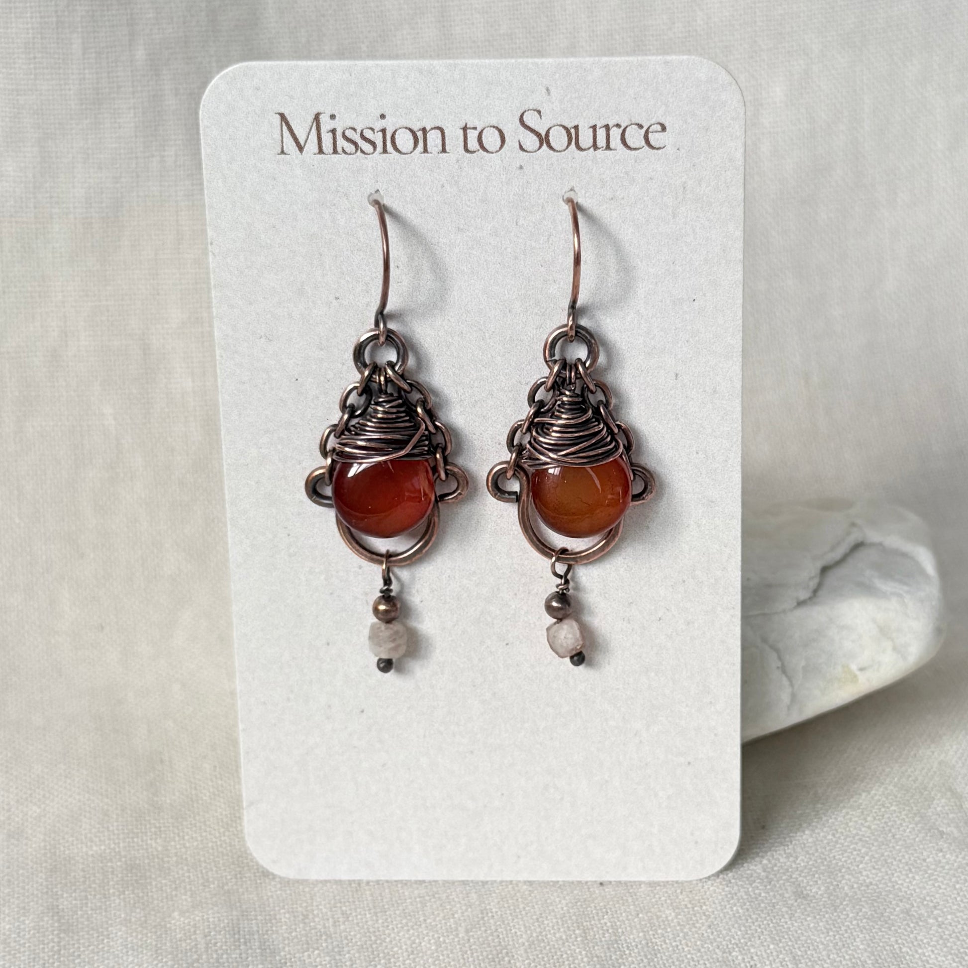 A pair of handmade wire-wrapped earrings with orange-red Carnelian stones and small white beads, displayed on a white card labeled 'Mission to Source' against a neutral fabric background.