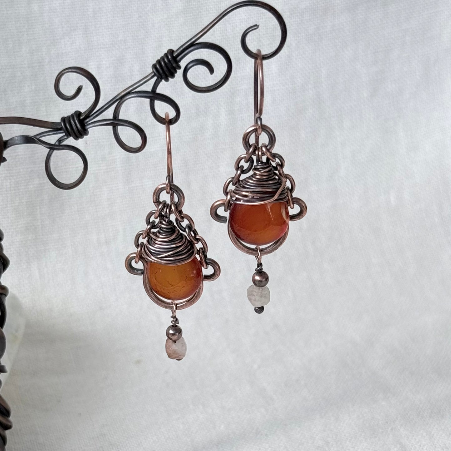 A pair of handmade wire-wrapped drop earrings featuring orange carnelian stones and small sunstone beads, hanging from a black metal display stand against a white fabric background.
