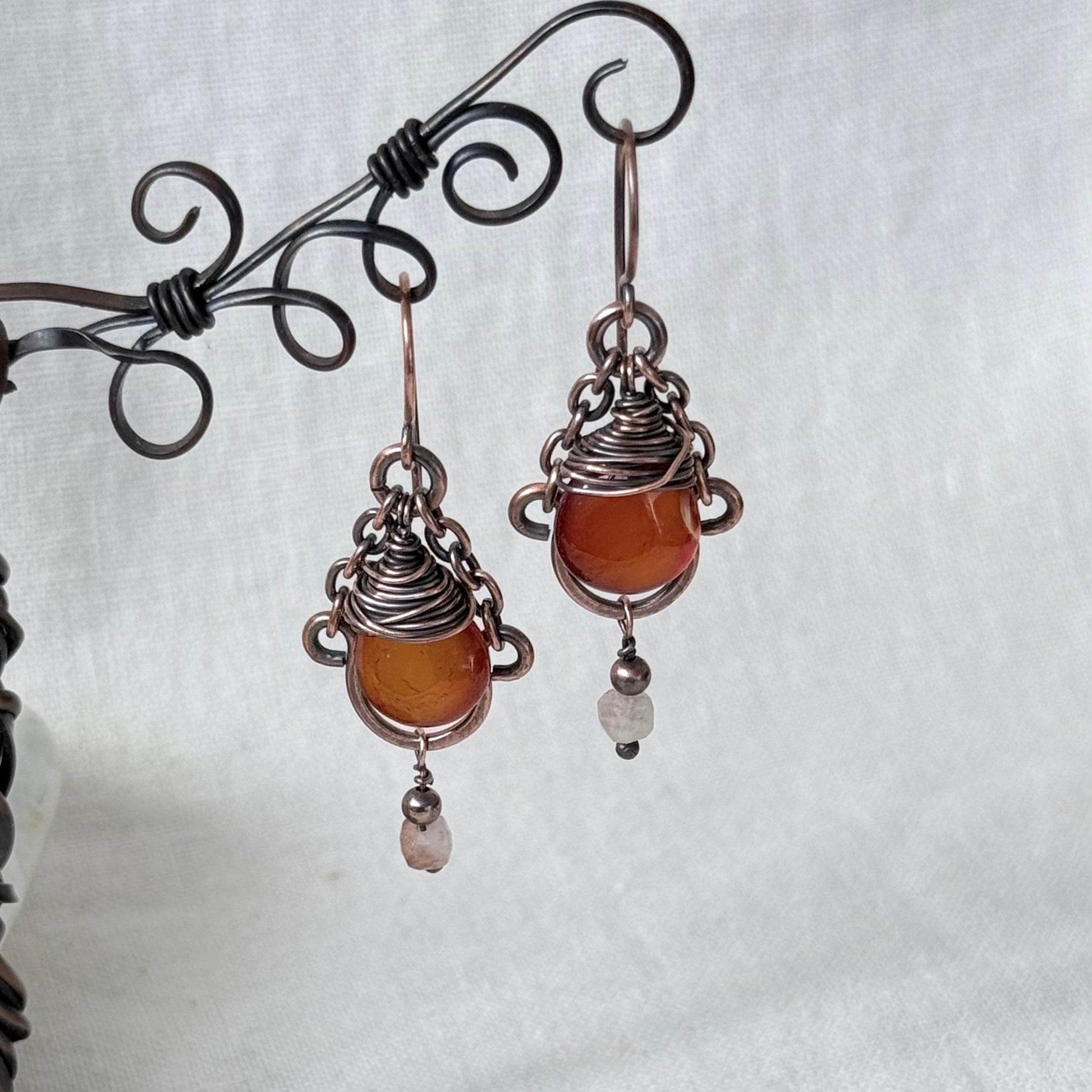 A pair of handmade wire-wrapped drop earrings featuring orange carnelian stones and small sunstone beads, hanging from a black metal display stand against a white fabric background.