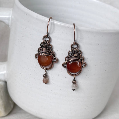 A pair of handmade drop earrings with copper wire wrapping and round orange-red carnelian stones, resting against a white speckled ceramic mug.