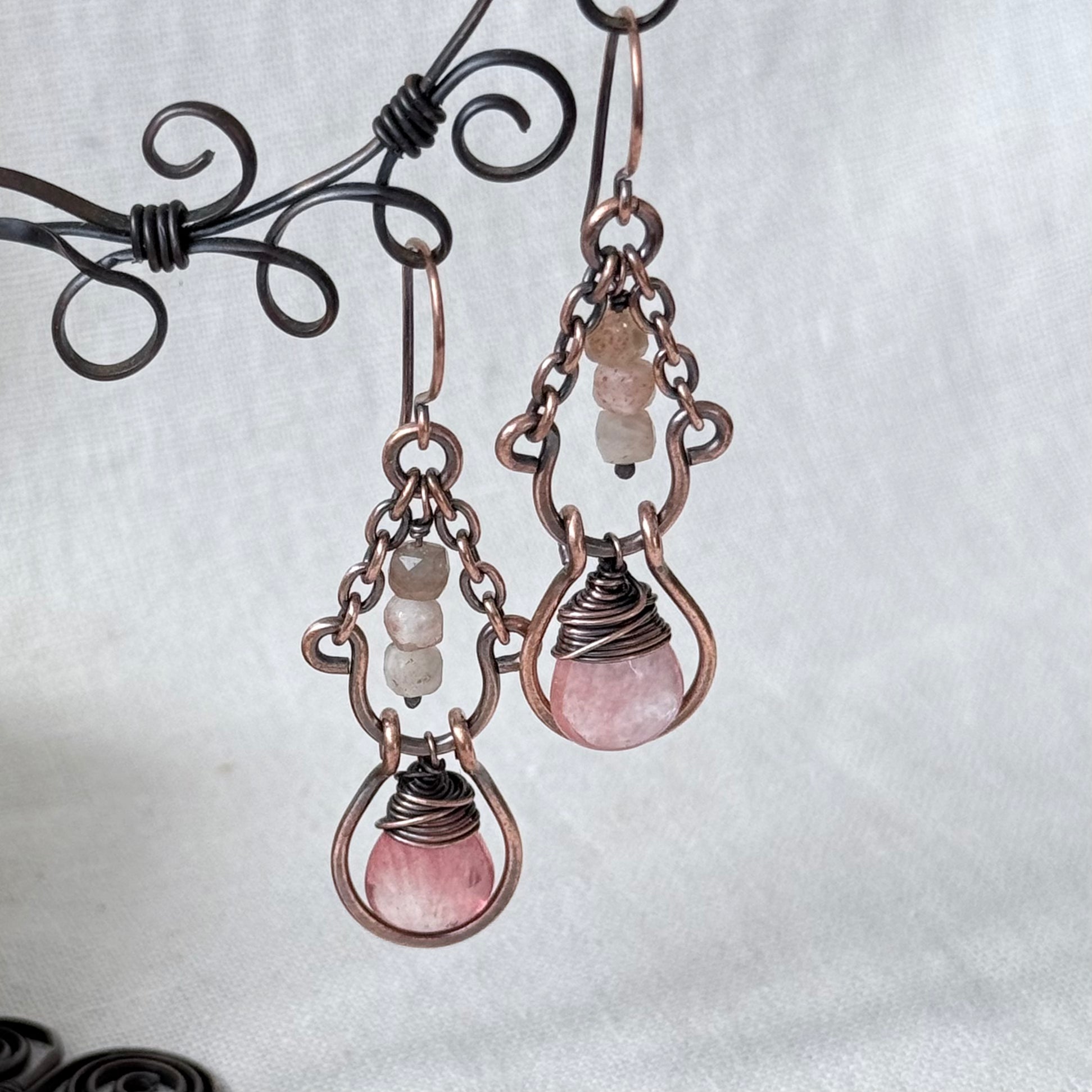 A pair of handmade copper wire drop earrings with pink and white beads, hanging from a black wire display stand against a white fabric background.