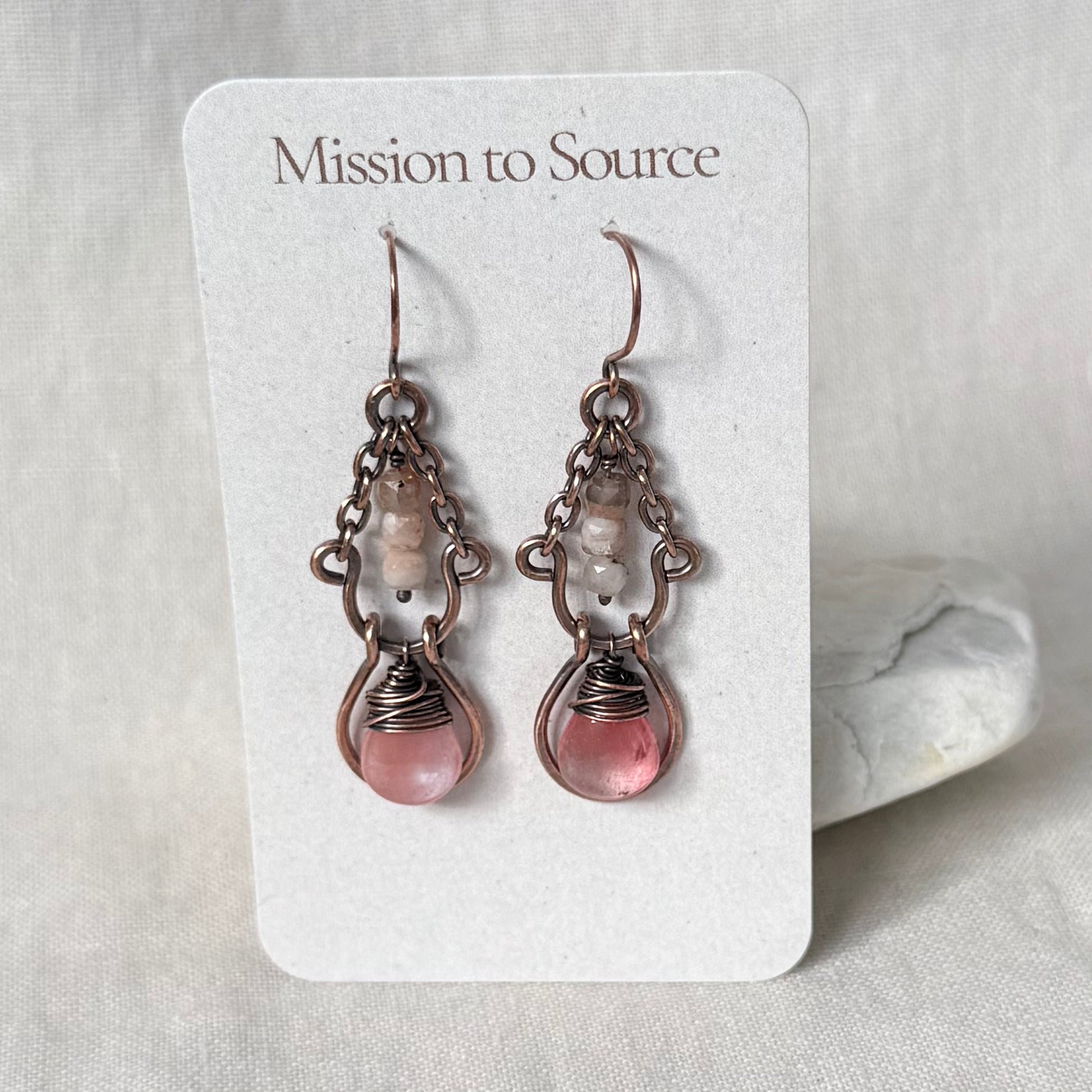 A pair of handmade dangle earrings displayed on a white card labeled 'Mission to Source.' The earrings have copper chains and frames with pale pink translucent beads and oval pink stone drops.