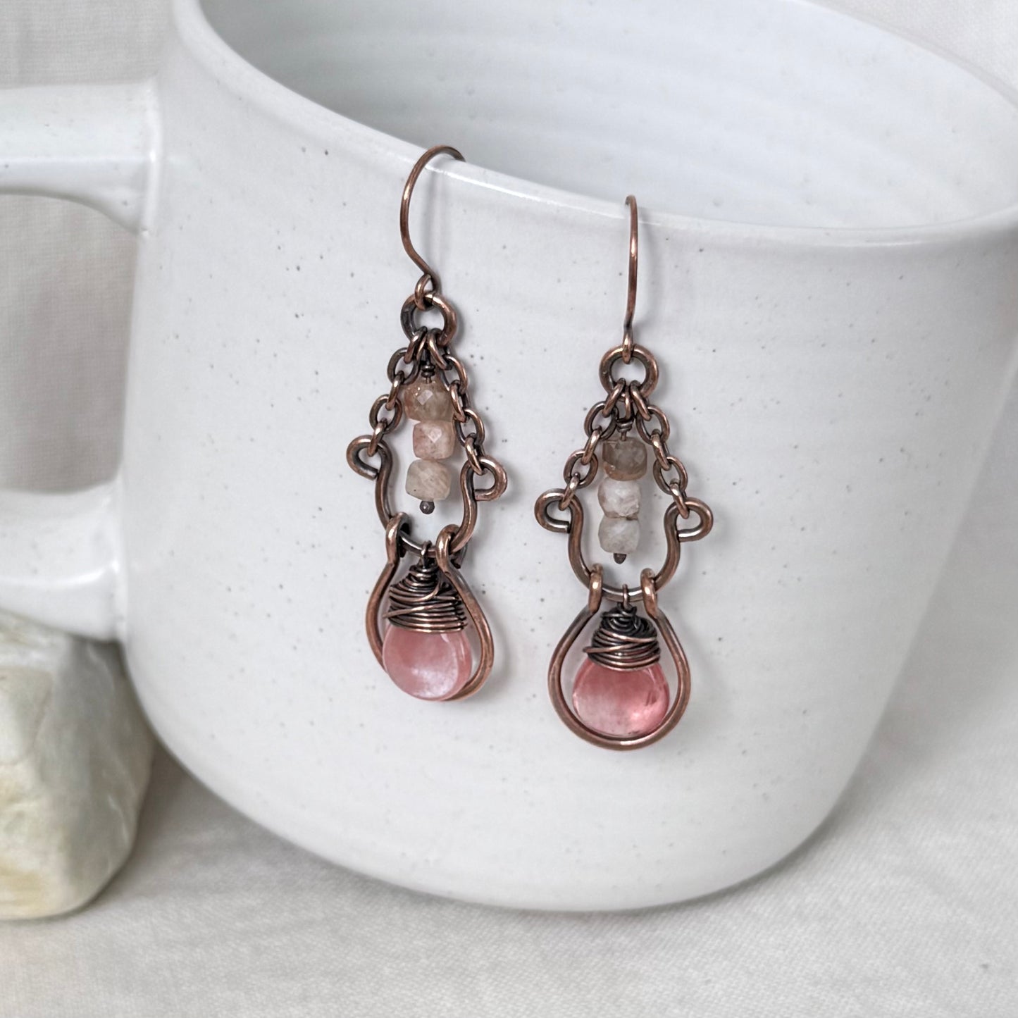 A pair of handmade drop earrings with copper metalwork, small clear beads, and pink teardrop-shaped stones, hanging against a white ceramic mug.