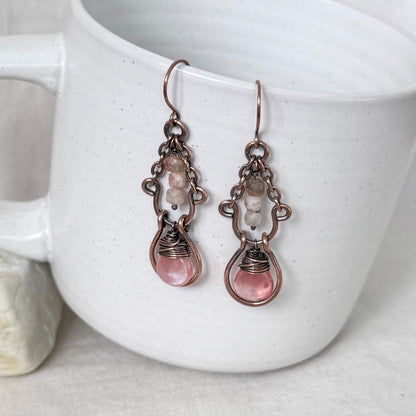 A pair of handmade drop earrings with copper metalwork, small clear beads, and pink teardrop-shaped stones, hanging against a white ceramic mug.