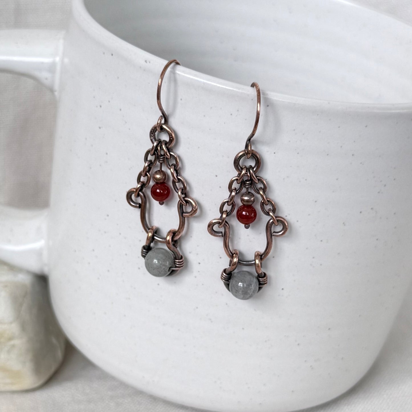 A pair of handmade copper dangle earrings, a small red bead, and a grey stone, hanging against a white speckled ceramic mug.