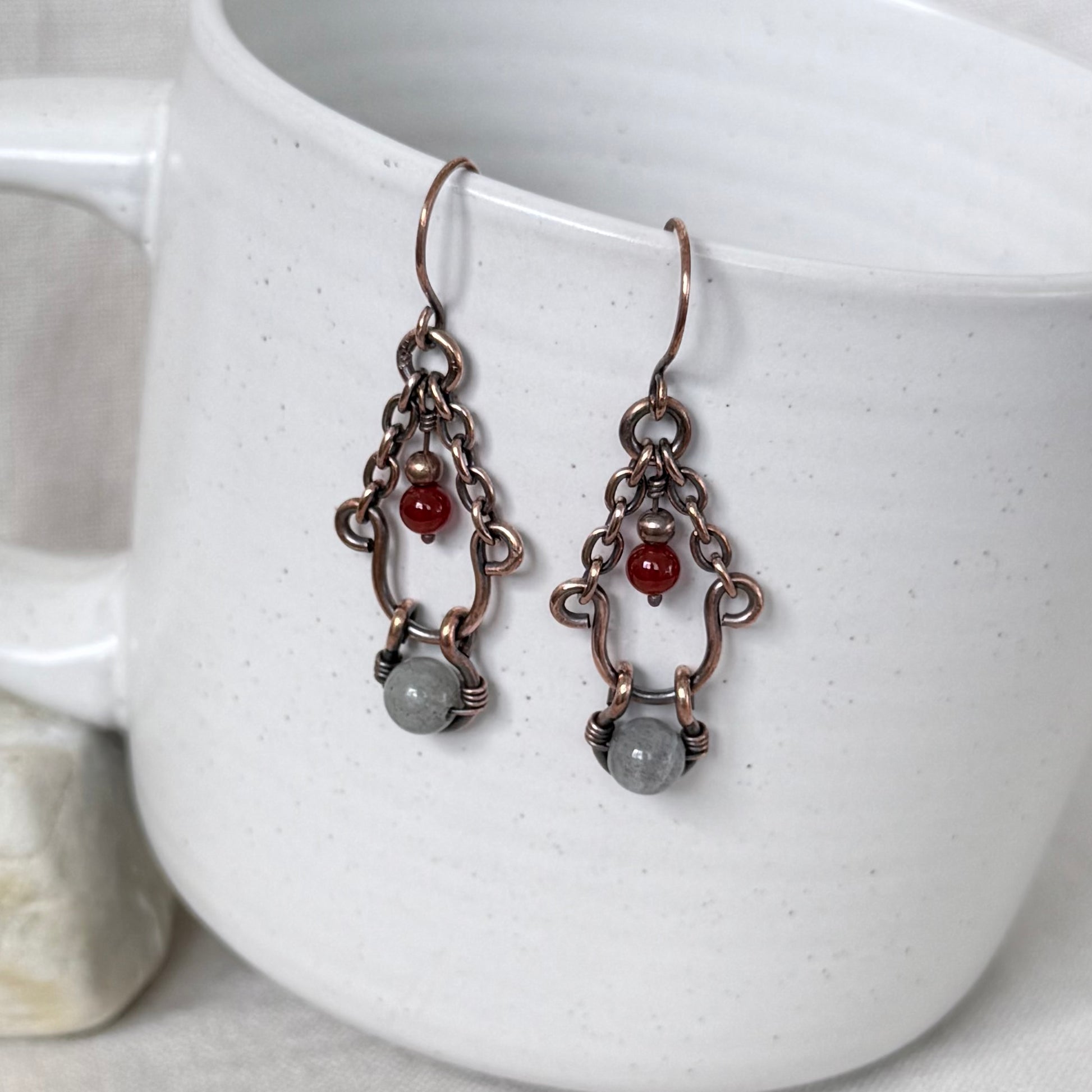 A pair of handmade copper dangle earrings, a small red bead, and a grey stone, hanging against a white speckled ceramic mug.
