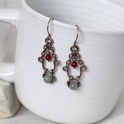 A pair of handmade copper dangle earrings, a small red bead, and a grey stone, hanging against a white speckled ceramic mug.