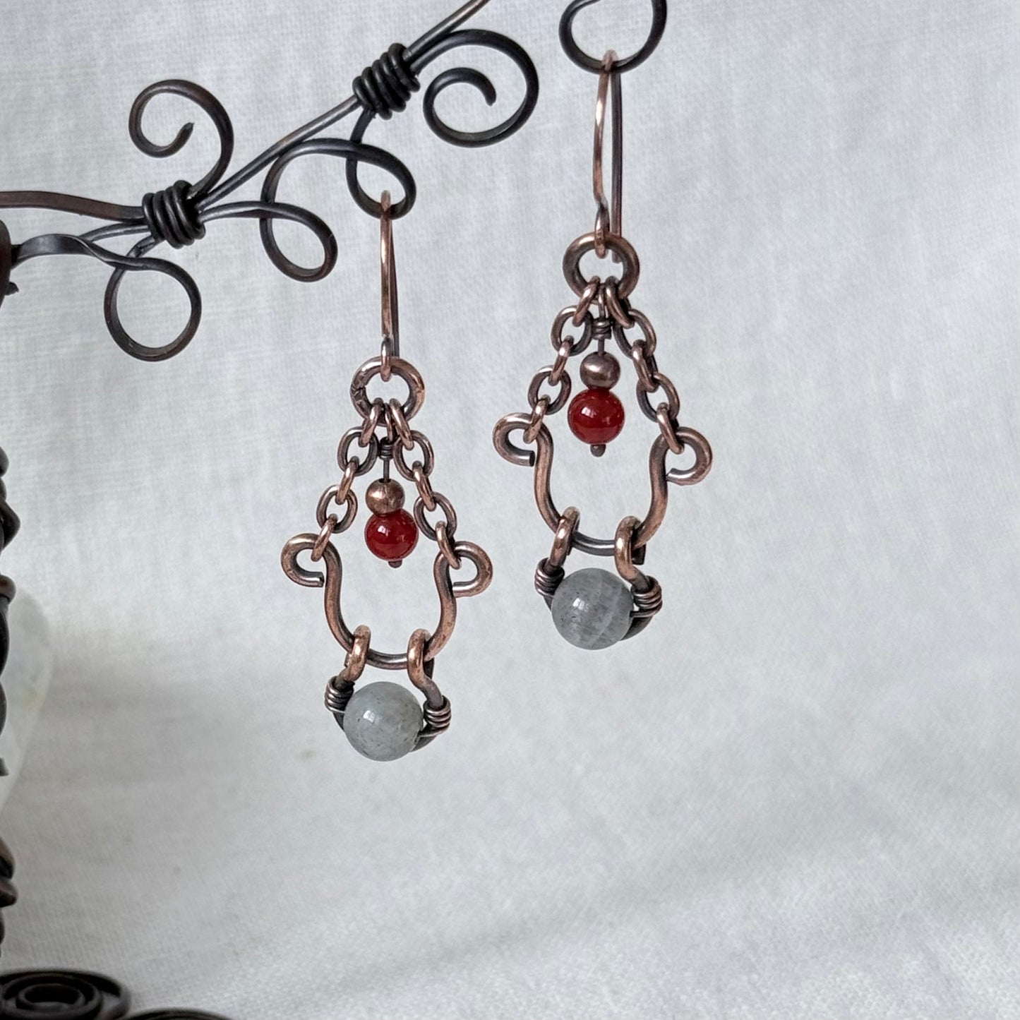 A pair of handmade copper wire dangle earrings with small red beads and gray stone charms, displayed on a black metal stand against a white fabric background.
