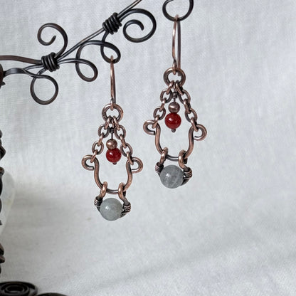 A pair of handmade copper wire dangle earrings with small red beads and gray stone charms, displayed on a black metal stand against a white fabric background.