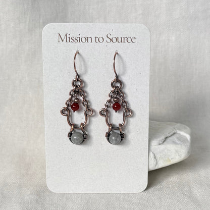 A pair of handmade dangle earrings displayed on a white card labeled 'Mission to Source.' The earrings have copper metalwork with small red beads and a larger pale grey stone at the bottom.