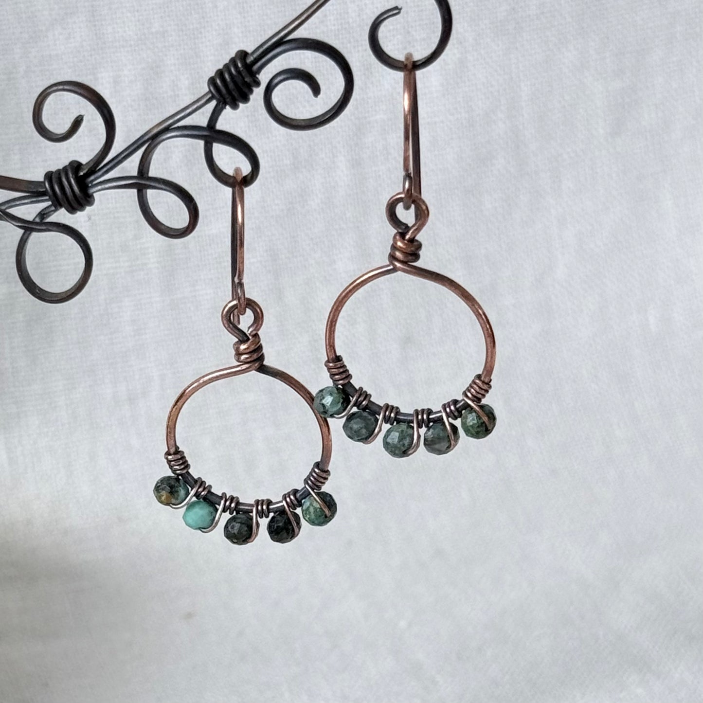 A pair of handmade copper wire hoop earrings with small green faceted beads, displayed on a black wire stand against a white textured fabric background.