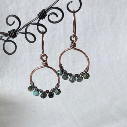 A pair of handmade copper wire hoop earrings with small green faceted beads, displayed on a black wire stand against a white textured fabric background.