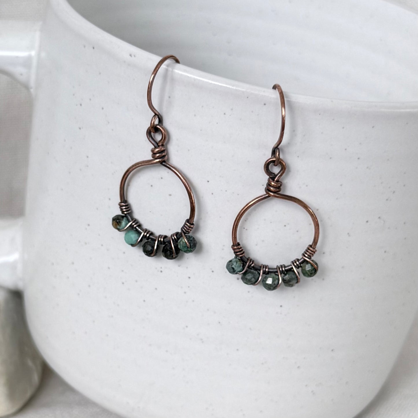 A pair of handmade copper wire hoop earrings with small green faceted beads, displayed hanging from the rim of a white speckled ceramic mug.