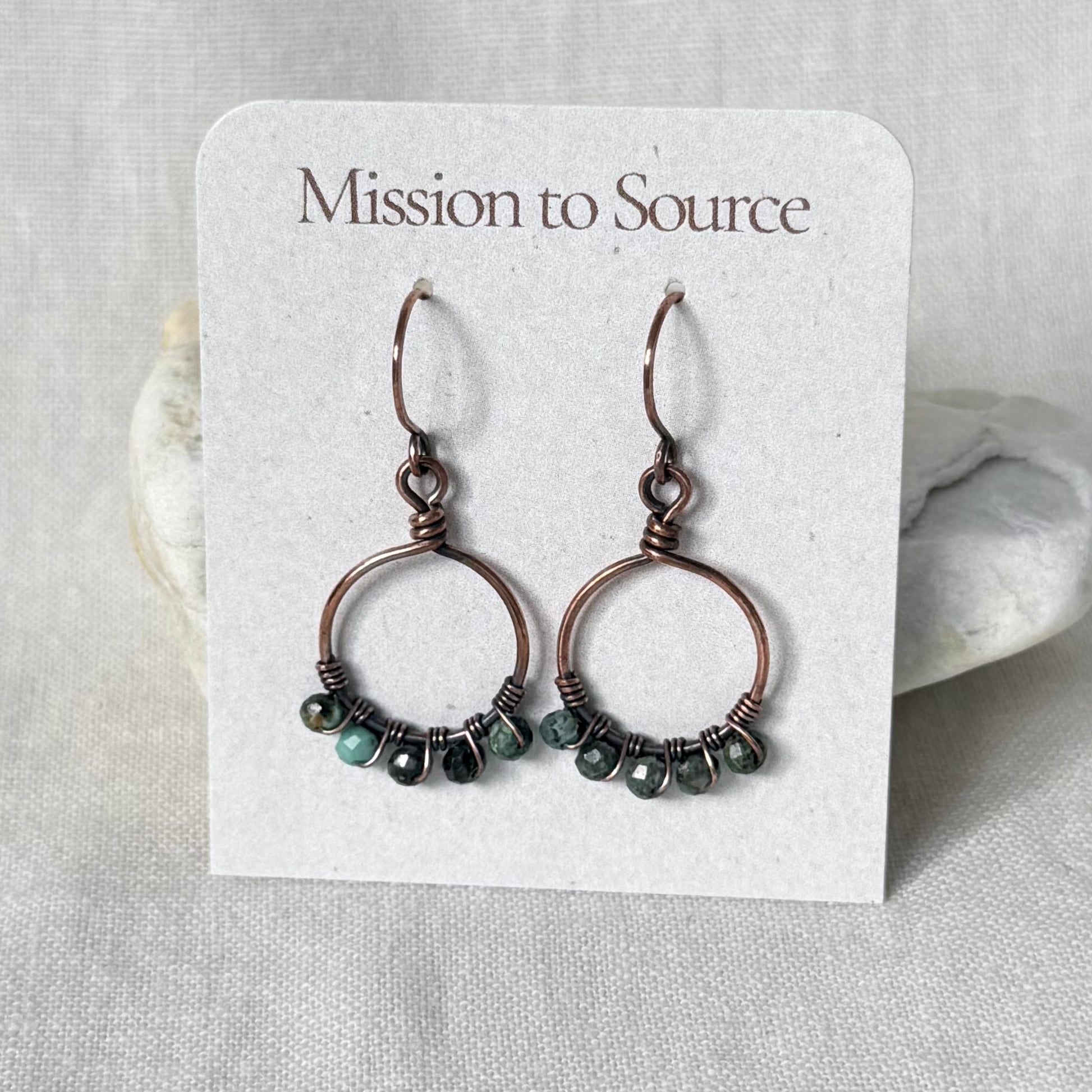A pair of handmade copper hoop earrings with small faceted dark green and blue beads, displayed on a white card labeled 'Mission to Source'.