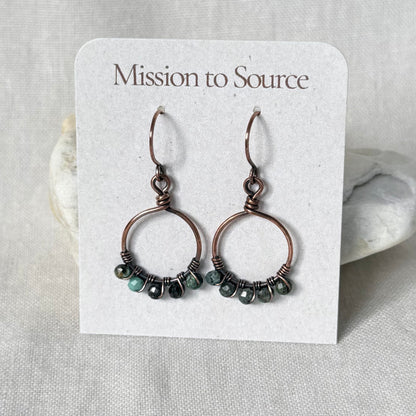 A pair of handmade copper hoop earrings with small faceted dark green and blue beads, displayed on a white card labeled 'Mission to Source'.