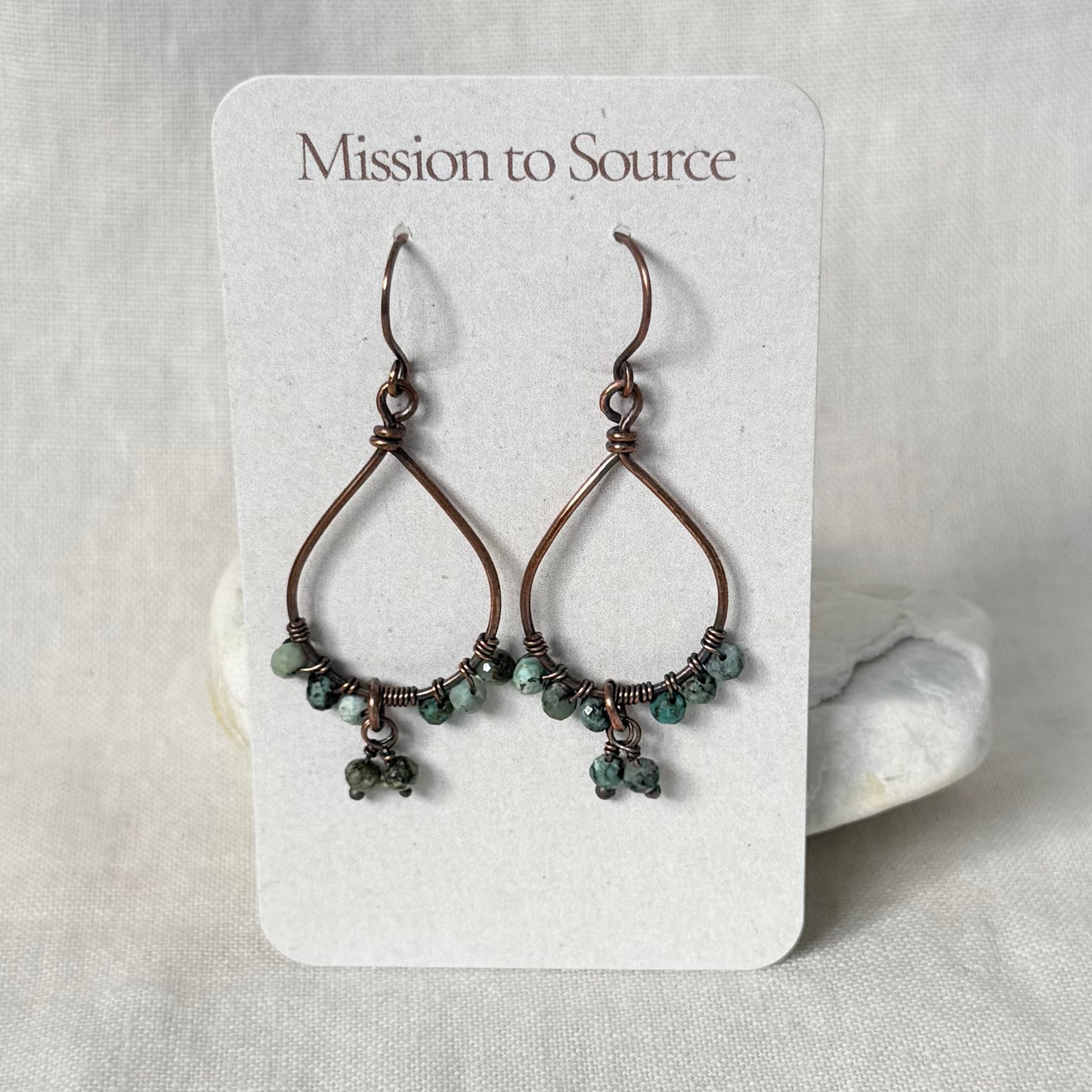 A pair of handmade teardrop-shaped wire earrings with small green beads, displayed on a white card labeled 'Mission to Source' against a neutral fabric background.