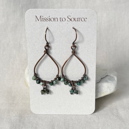 A pair of handmade teardrop-shaped wire earrings with small green beads, displayed on a white card labeled 'Mission to Source' against a neutral fabric background.