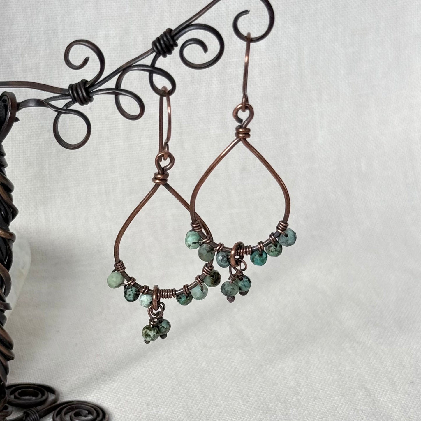 Handmade copper wire dangle earrings with small green faceted beads, displayed on a black metal stand against a white fabric background.