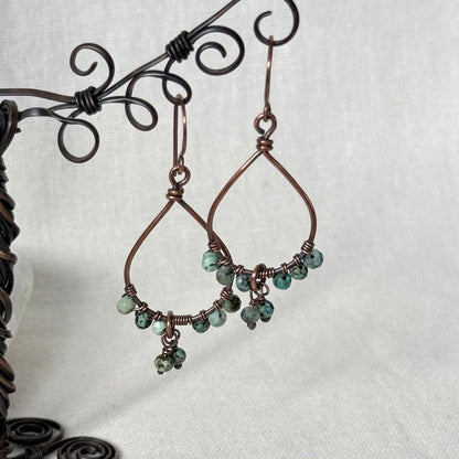 Handmade copper wire dangle earrings with small green faceted beads, displayed on a black metal stand against a white fabric background.