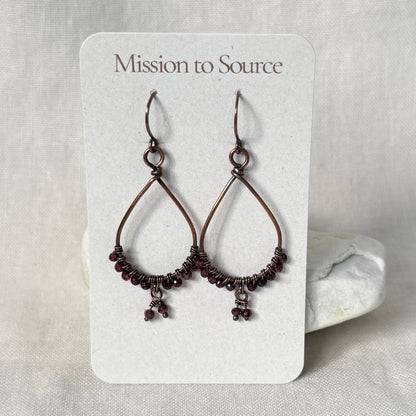 A pair of handmade teardrop dangle earrings with dark copper wire frames, wrapped beads, and small dark purple beads hanging from the bottom, displayed on a white card labeled 'Mission to Source'.
