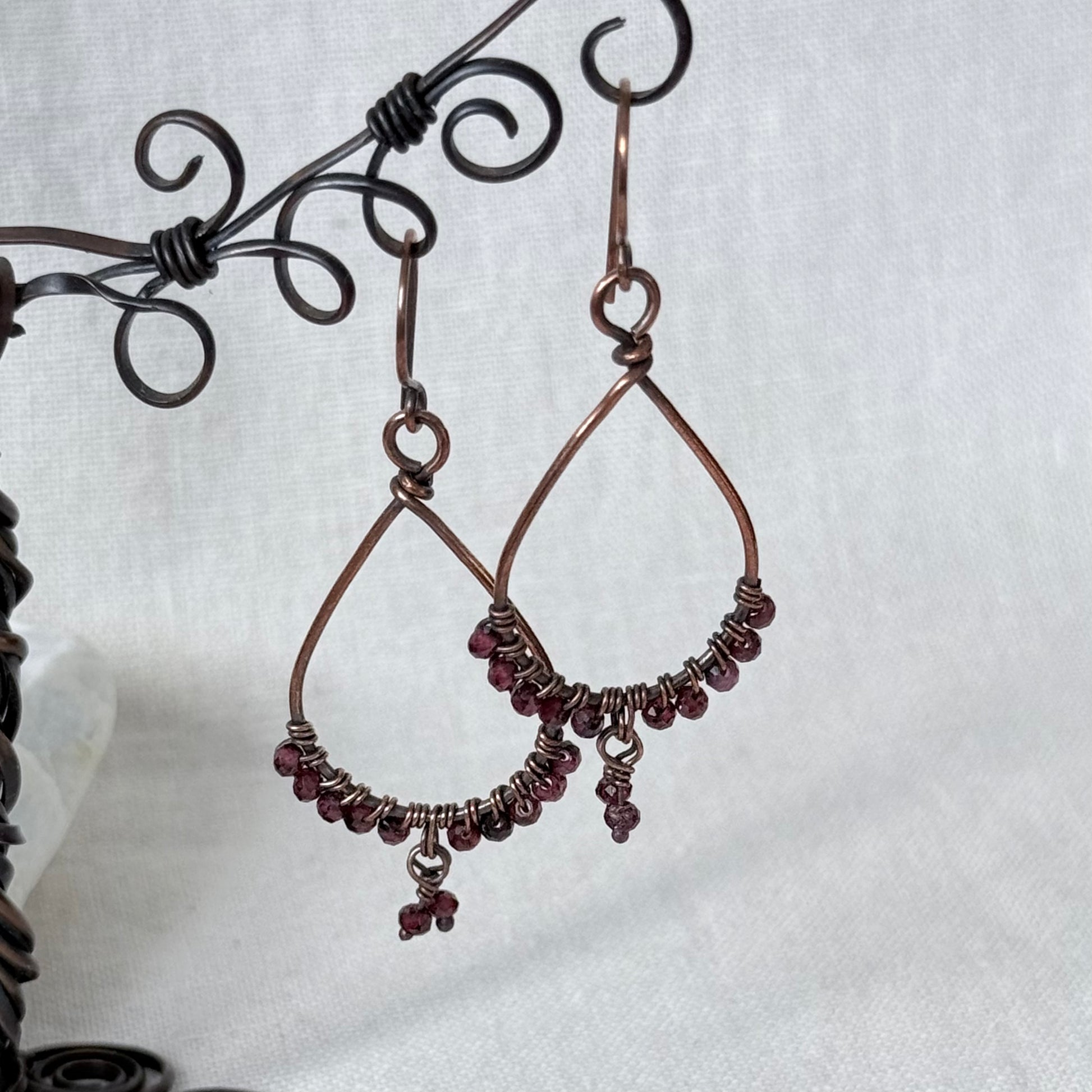 A pair of handmade copper wire teardrop dangle earrings with small dark red beads along the bottom curve, hanging from a black metal wire display stand against a light gray fabric background.