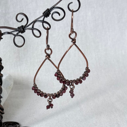 A pair of handmade copper wire teardrop dangle earrings with small dark red beads along the bottom curve, hanging from a black metal wire display stand against a light gray fabric background.