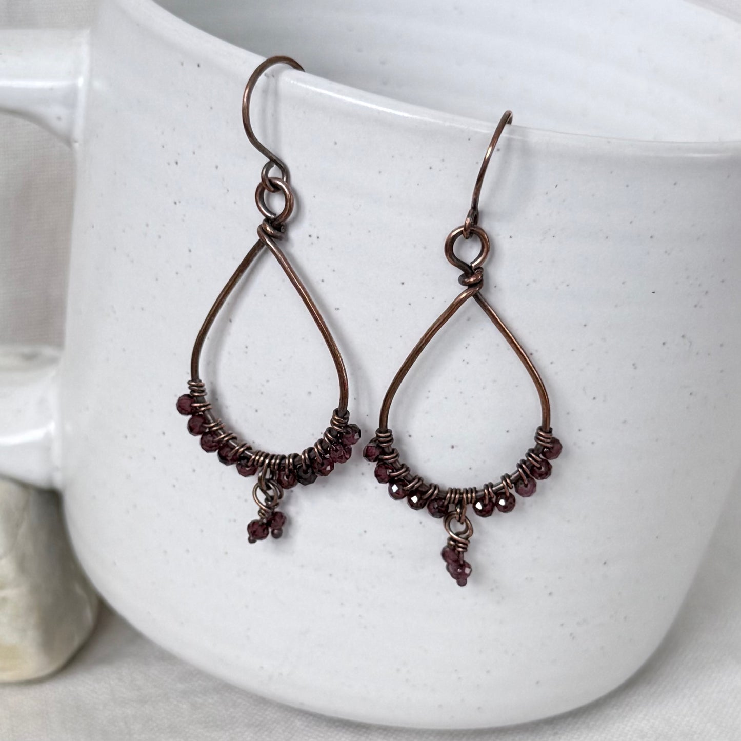 A pair of handmade teardrop-shaped dangle earrings with dark copper wire frames, wrapped bead accents, and small dangling beads, displayed against a white speckled ceramic mug.