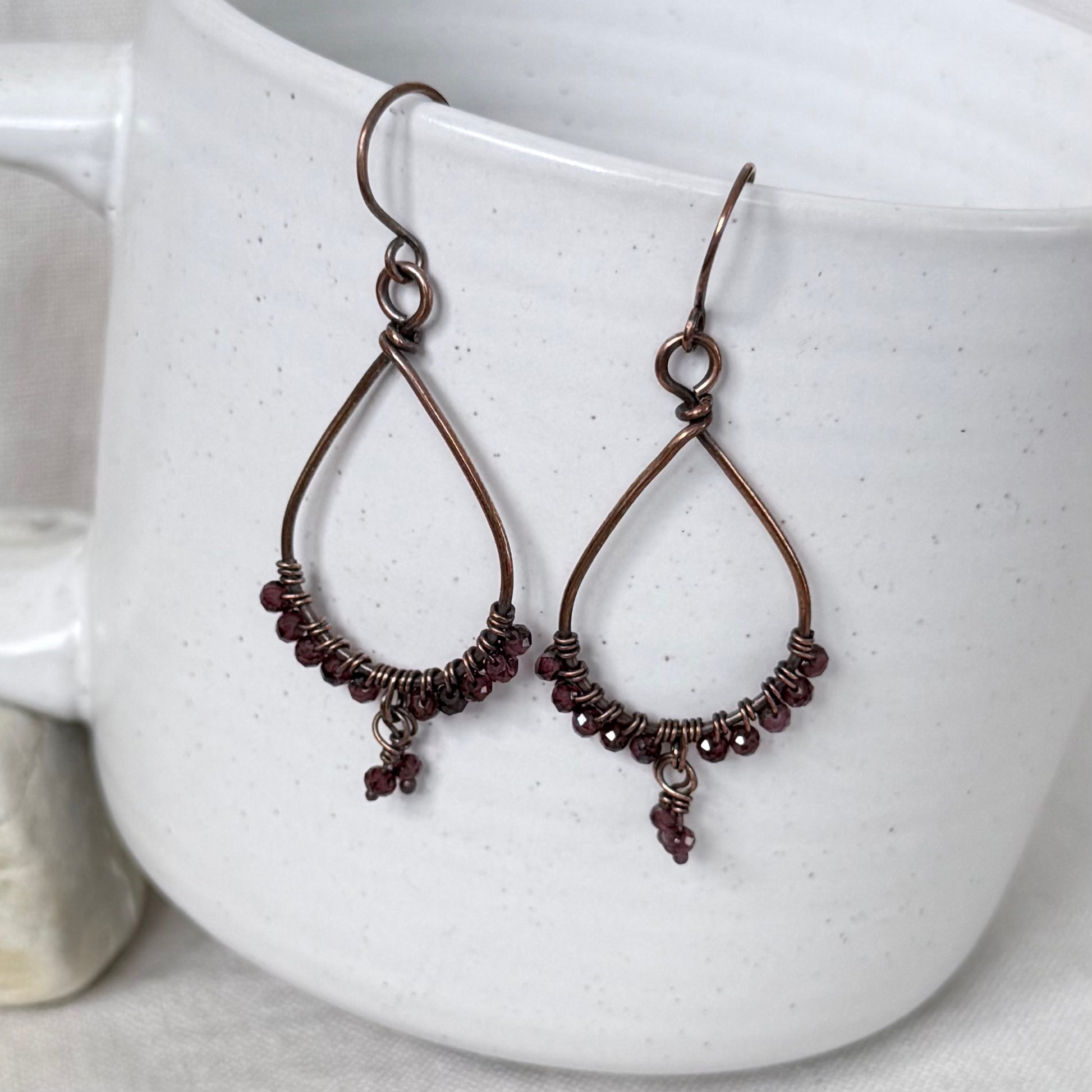 A pair of handmade teardrop-shaped dangle earrings with dark copper wire frames, wrapped bead accents, and small dangling beads, displayed against a white speckled ceramic mug.