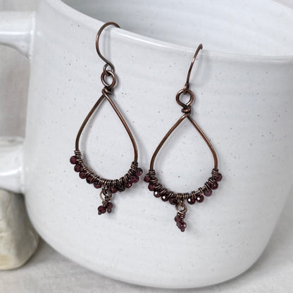 A pair of handmade teardrop-shaped dangle earrings with dark copper wire frames, wrapped bead accents, and small dangling beads, displayed against a white speckled ceramic mug.