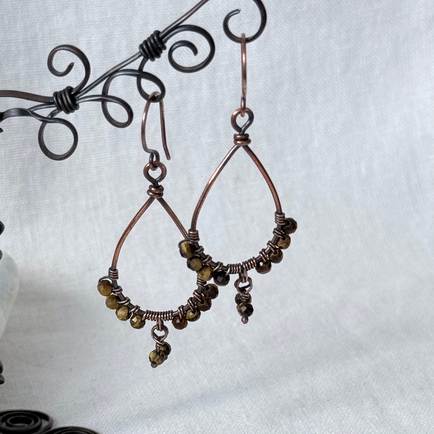 A pair of handmade copper wire hoop earrings with small dark brown beads and dangling charms, displayed on a black scroll stand against a light gray fabric background.