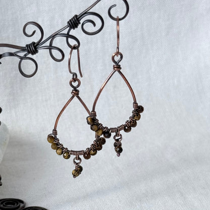 A pair of handmade copper wire hoop earrings with small dark brown beads and dangling charms, displayed on a black scroll stand against a light gray fabric background.