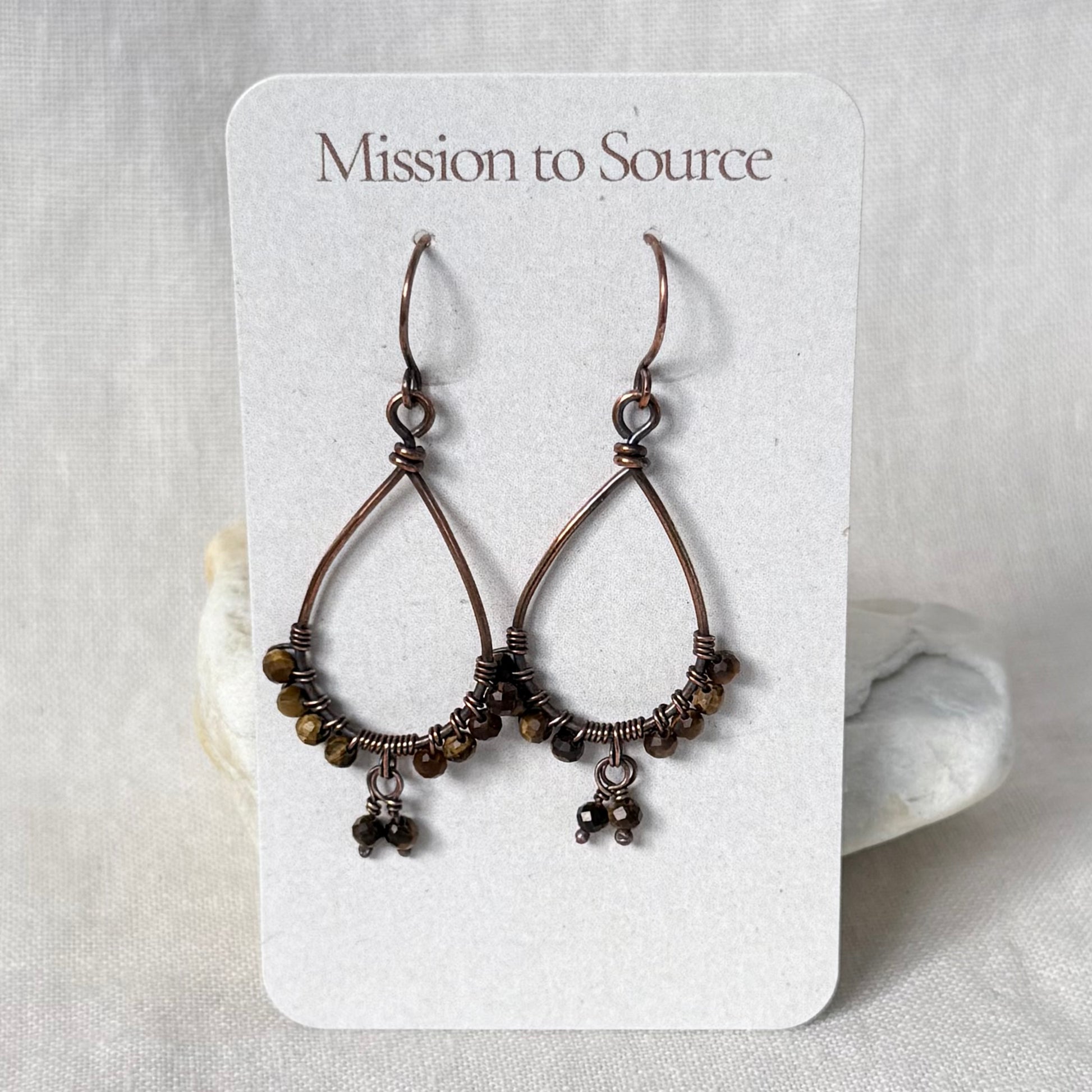 A pair of copper dangle earrings with small brown beads and dangling charms, displayed on a white card labeled 'Mission to Source' against a light fabric background.