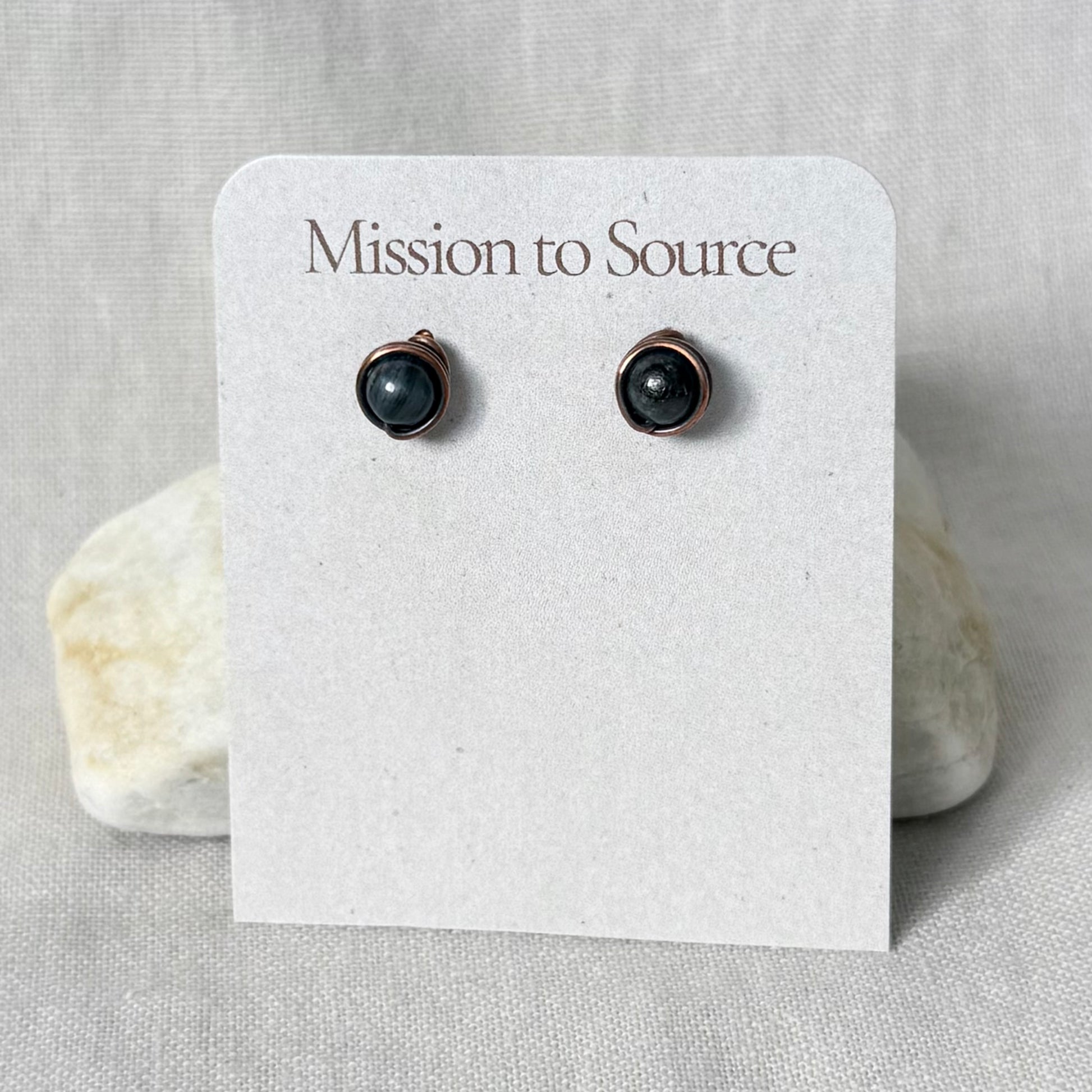 A pair of small round copper wire stud earrings with dark Apatite beads, displayed on a white card that reads 'Mission to Source' against a neutral textured background.