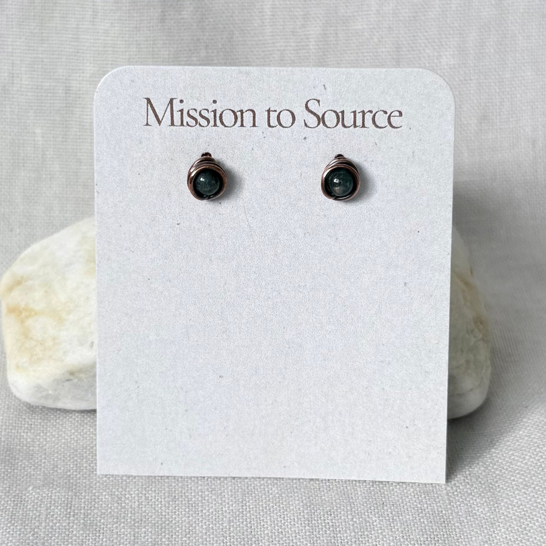 A pair of small dark green gemstone stud earrings in a copper wire wrap setting, displayed on a white paper card that reads 'Mission to Source.' The card is photographed against a light neutral fabric background with two small stones behind it.