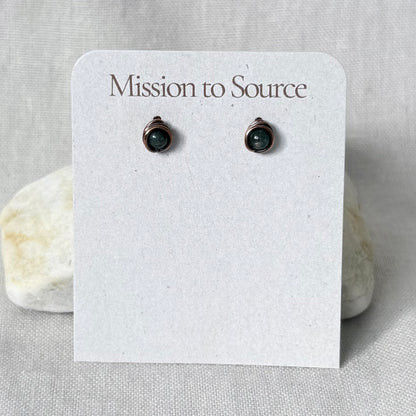 A pair of small dark green gemstone stud earrings in a copper wire wrap setting, displayed on a white paper card that reads 'Mission to Source.' The card is photographed against a light neutral fabric background with two small stones behind it.
