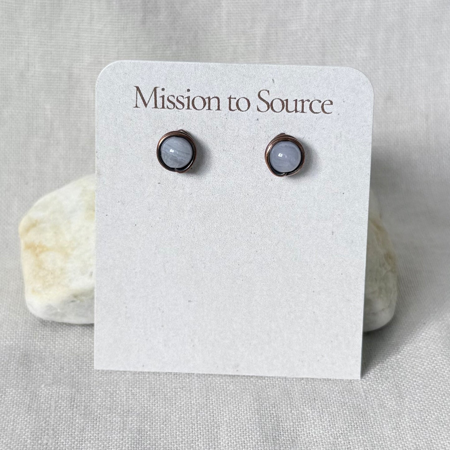 A pair of round stud earrings with pale gray-blue stones in dark copper settings, displayed on a white card printed with the brand name 'Mission to Source.' The card is photographed against a light neutral fabric background with two small stones behind it.