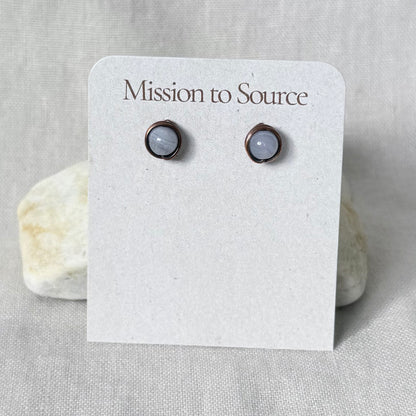 A pair of round stud earrings with pale gray-blue stones in dark copper settings, displayed on a white card printed with the brand name 'Mission to Source.' The card is photographed against a light neutral fabric background with two small stones behind it.