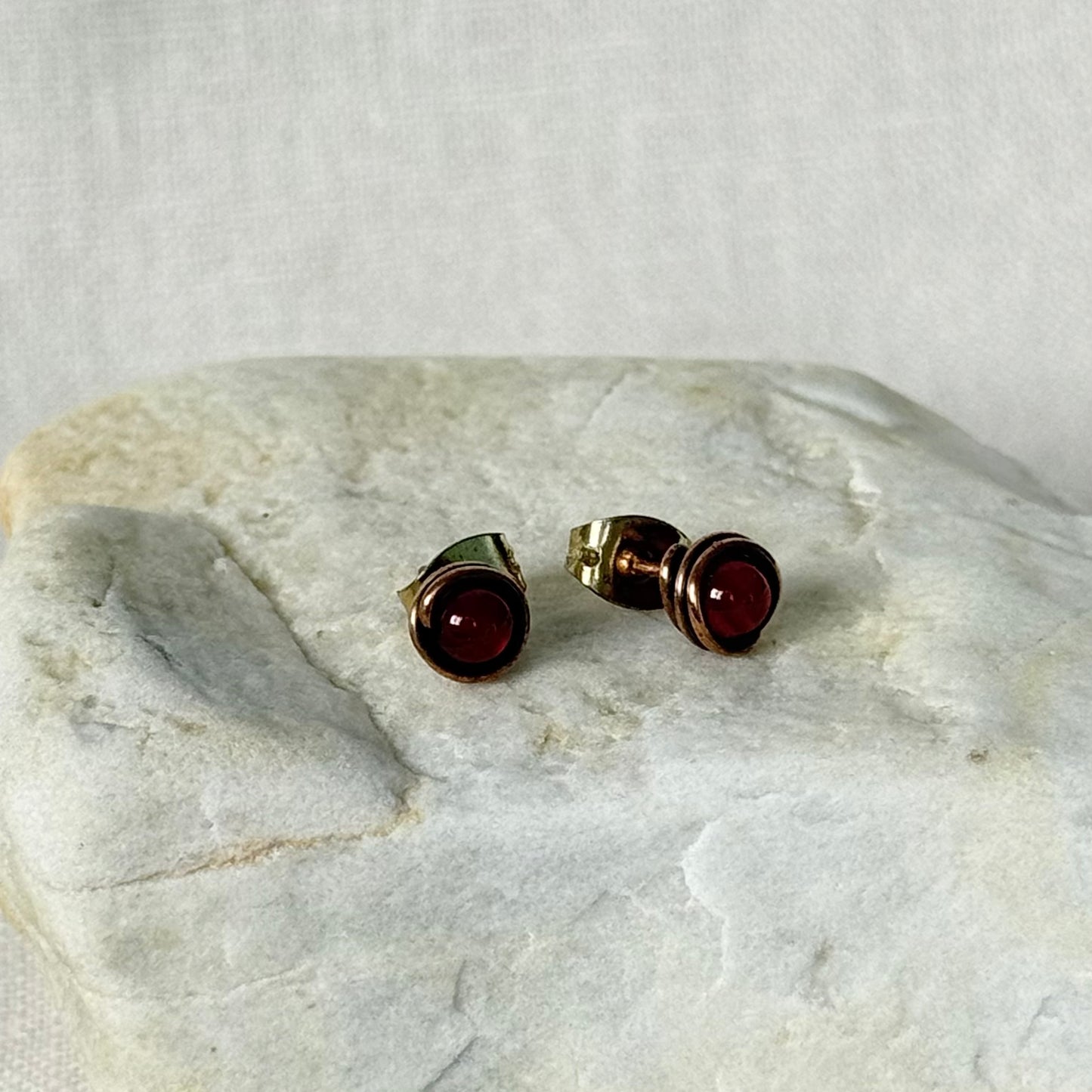 A pair of copper stud earrings with round dark red gemstones, displayed on a textured light grey stone slab against a neutral fabric background.