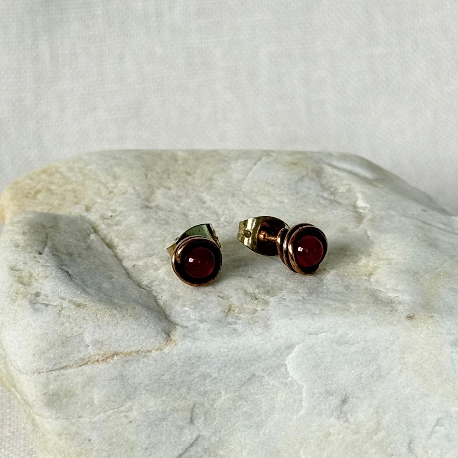 A pair of copper stud earrings with round dark red gemstones, displayed on a textured light grey stone slab against a neutral fabric background.