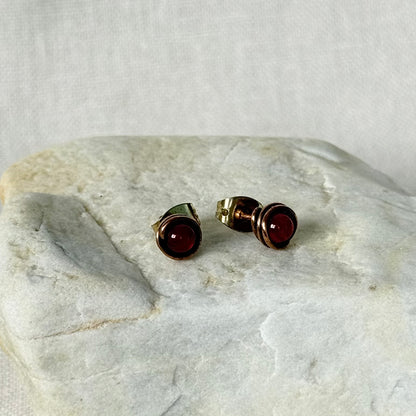 A pair of copper stud earrings with round dark red gemstones, displayed on a textured light grey stone slab against a neutral fabric background.