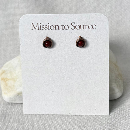 A pair of small wrap-style stud earrings with dark red round stones in a wire setting, displayed on a white card that reads "Mission to Source," set against a light fabric background with two small stones behind the card.