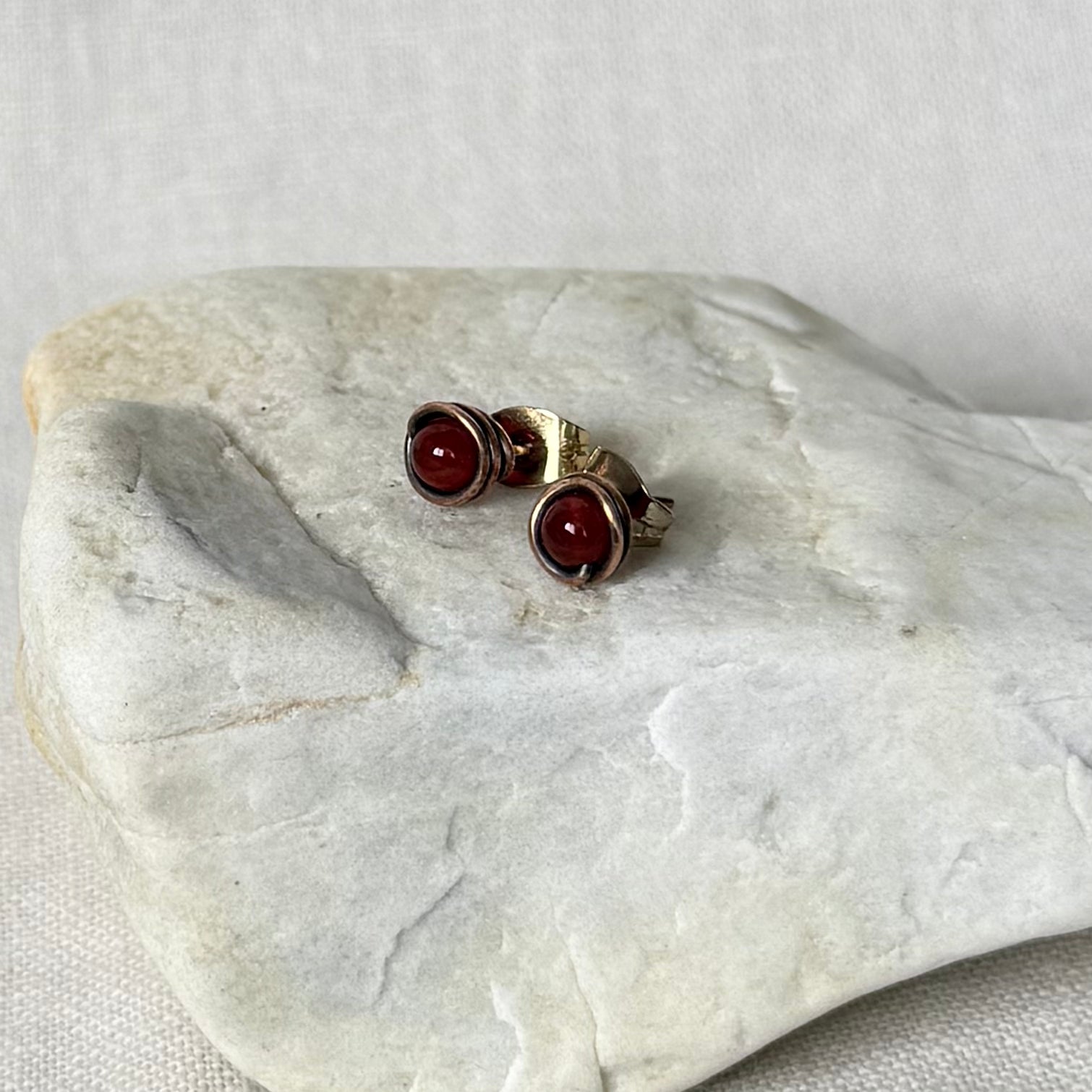 A pair of copper stud earrings with round dark red gemstones, displayed on a light grey stone slab against a neutral fabric background.