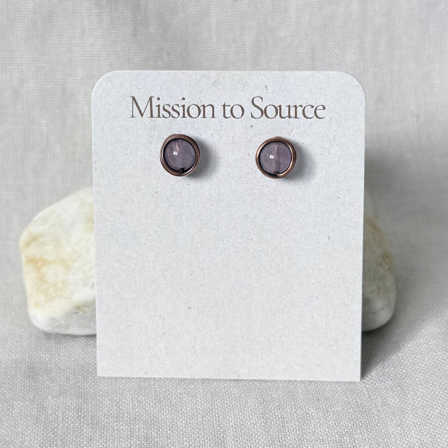 A pair of small round stud earrings with light purple stones in a copper wire wrap setting, displayed on a white card labeled "Mission to Source" against a light fabric background with two stones behind it.