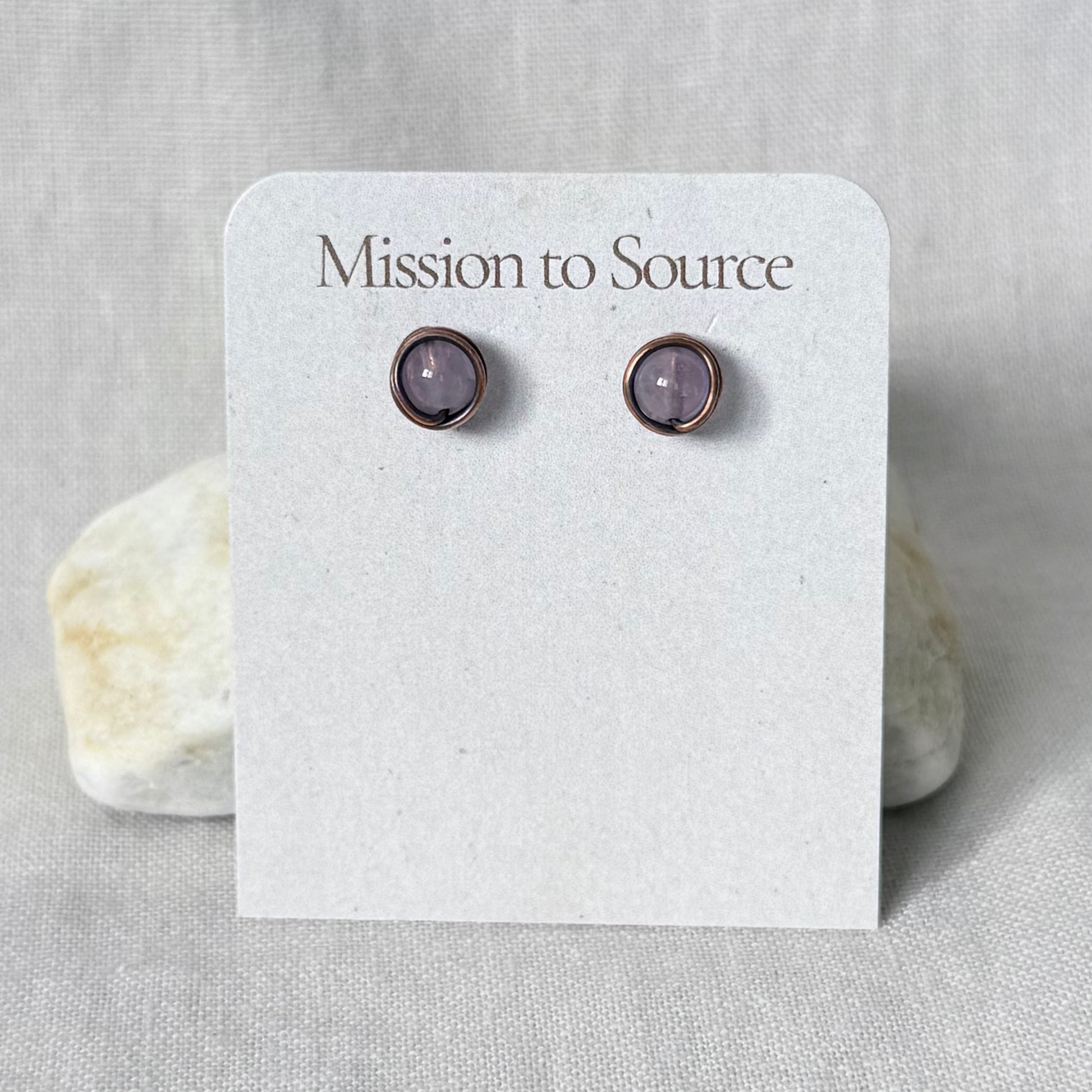 A pair of small round stud earrings with light purple stones in a copper wire wrap setting, displayed on a white card labeled "Mission to Source" against a light fabric background with two stones behind it.